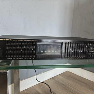 Può includere: Equalizzatore grafico Marantz nero. Il componente audio presenta numerosi cursori, uno schermo e varie manopole e pulsanti di controllo. L'unità è su una mensola di vetro.