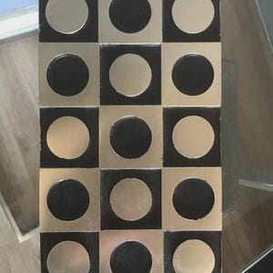 Może przedstawiać: Prostokątny przedmiot dekoracyjny z wzorem szachownicy. Projekt przedstawia naprzemiennie srebrne koła i czarne kwadraty. Obiekt jest wykonany z metalu i ma odblaskową powierzchnię.