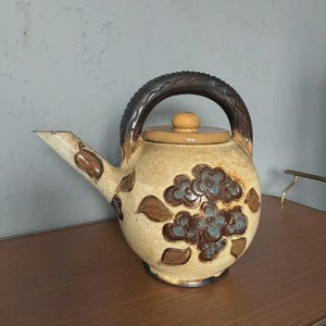 Op de afbeelding: Een keramische theepot met een bruine handgreep en deksel. De beige body is versierd met bruine en blauwe bloemmotieven. De tuit en handgreep hebben een textuur. Een keukengerei in vintage-stijl.