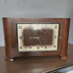 Op de afbeelding: Een vintage houten klok met een rechthoekige wijzerplaat en een donkerbruine afwerking. De wijzerplaat heeft zwarte cijfers en wijzers, met een glazen afdekking. De klok staat op een houten oppervlak.