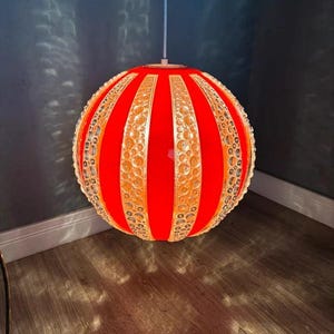 May include: A red and gold pendant light fixture. The spherical lamp has alternating vertical panels of red and gold, with the gold panels featuring a decorative, openwork design. The lamp hangs from a white cord.