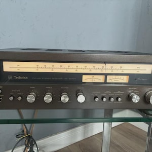 Puede incluir: Un receptor estéreo FM/AM Technics SA-505X vintage. El receptor marrón oscuro tiene una pantalla con una escala de frecuencia y múltiples diales y botones para volumen, balance y sintonización. El logotipo de Technics es visible.