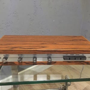 May include: A vintage stereo receiver with a wood grain effect. The silver faceplate features control knobs for volume, balance, bass, and treble. It has buttons for radio functions. The brand name "LUXOR" is displayed on the right.