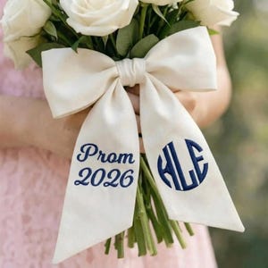 May include: A bouquet of white roses is decorated with a large cream-coloured bow. The bow has navy blue embroidered text reading "Prom 2026" and a monogram. The flowers and bow are held against a soft pink background.