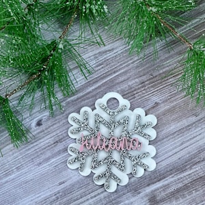 K&ouml;nnte beinhalten: Ein wei&szlig;er Schneeflocken-Anh&auml;nger mit silbernem Glitzer und dem Namen "Juliana" in rosafarbenem Glitzer.