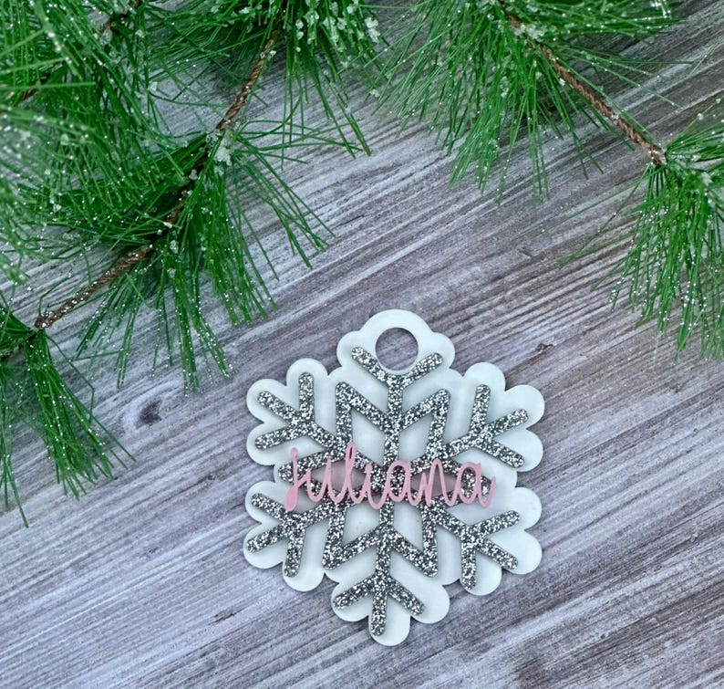 K&ouml;nnte beinhalten: Ein wei&szlig;er Schneeflocken-Anh&auml;nger mit silbernem Glitzer und dem Namen "Juliana" in rosafarbenem Glitzer.