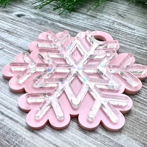 K&ouml;nnte beinhalten: Ein rosa Schneeflocken-Anh&auml;nger mit einem klaren, glitzernden Schneeflocken-Design im Inneren. Der Anh&auml;nger ist aus Harz gefertigt und hat oben ein Loch zum Aufh&auml;ngen.