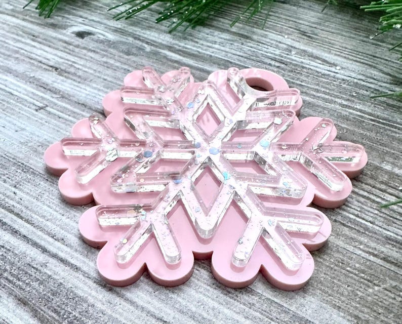 K&ouml;nnte beinhalten: Ein rosa Schneeflocken-Anh&auml;nger mit einem klaren, glitzernden Schneeflocken-Design im Inneren. Der Anh&auml;nger ist aus Harz gefertigt und hat oben ein Loch zum Aufh&auml;ngen.