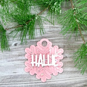 K&ouml;nnte beinhalten: Rosaer Schneeflocken-Anh&auml;nger mit Glitzer und dem Namen "Hallie" in wei&szlig;.