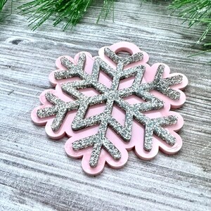 K&ouml;nnte beinhalten: Ein rosa Schneeflocken-Anh&auml;nger mit silbernem Glitzerfinish. Die Schneeflocke hat oben ein Loch zum Aufh&auml;ngen.