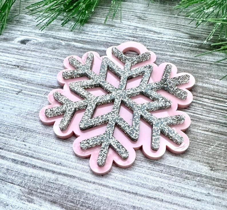 K&ouml;nnte beinhalten: Ein rosa Schneeflocken-Anh&auml;nger mit silbernem Glitzerfinish. Die Schneeflocke hat oben ein Loch zum Aufh&auml;ngen.