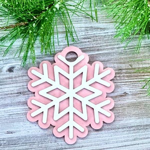 K&ouml;nnte beinhalten: Ein rosa Schneeflocken-Anh&auml;nger mit einem wei&szlig;en Schneeflocken-Design in der Mitte. Der Anh&auml;nger hat oben ein Loch zum Aufh&auml;ngen.