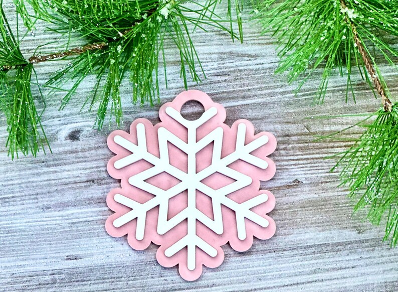 K&ouml;nnte beinhalten: Ein rosa Schneeflocken-Anh&auml;nger mit einem wei&szlig;en Schneeflocken-Design in der Mitte. Der Anh&auml;nger hat oben ein Loch zum Aufh&auml;ngen.