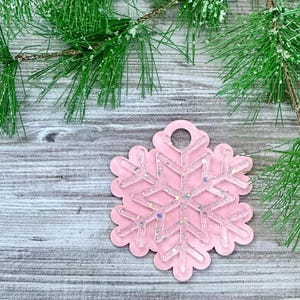 K&ouml;nnte beinhalten: Ein rosa Schneeflocken-Anh&auml;nger mit Glitzerakzenten, der an einem rustikalen Holzgrund mit einem Kiefernzweig h&auml;ngt.