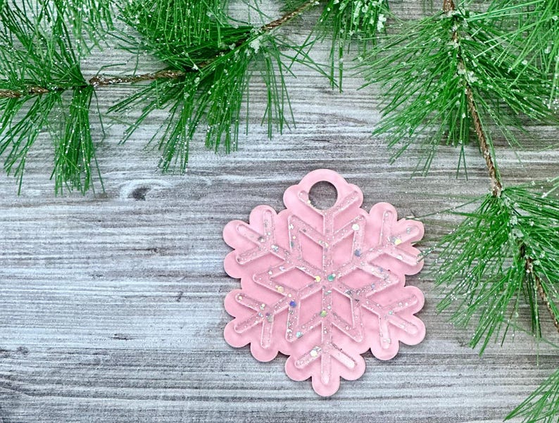 K&ouml;nnte beinhalten: Ein rosa Schneeflocken-Anh&auml;nger mit Glitzerakzenten, der an einem rustikalen Holzgrund mit einem Kiefernzweig h&auml;ngt.