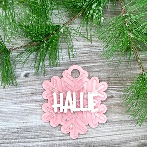 K&ouml;nnte beinhalten: Rosaer Schneeflocken-Anh&auml;nger mit Glitzer und dem Namen "Hallie" in wei&szlig;.