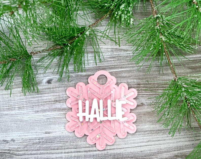 K&ouml;nnte beinhalten: Rosaer Schneeflocken-Anh&auml;nger mit Glitzer und dem Namen "Hallie" in wei&szlig;.