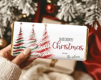 May include: A white chocolate bar wrapper with gold edges, featuring three Christmas trees in red and green. The words "Merry Christmas" are written in a festive font, with "from the wilsons" below.