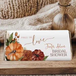 May include: A rectangular candy bar wrapper featuring a white background with watercolor illustrations of orange and dark red pumpkins, green leaves, and berries. The text reads FALL IN love, Jennifer Adams, and BRIDAL SHOWER in brown script and print.
