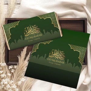 Könnte beinhalten: Zwei Schokoriegelverpackungen mit grün-goldenen Design, arabischer Kalligraphie und dem Text "EID MUBARAK". Die Verpackungen liegen auf einem Holztablett mit dekorativen Elementen.
