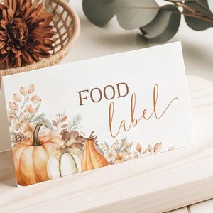 以下が含まれることがあります： 茶色の筆記体で「FOOD label」と書かれた折り畳まれたフードラベル。オレンジ、白、茶色の色合いでカボチャと秋の葉のイラストが描かれています。ラベルは明るい木製の表面にあります。