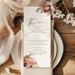 May include: A wedding menu card with floral accents, featuring a menu for a wedding. The menu includes a starter salad, beef fillet, and wedding cake. The card is on a white plate with a beige napkin, with flowers and a wooden background.