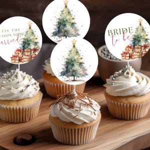 Puede incluir: Cupcakes con glaseado blanco y adornos decorativos con árboles de Navidad y texto. Un adorno dice "Tis the season to be married", y otro dice "Bride to be". Los cupcakes están sobre una tabla de madera.