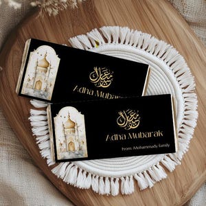 Peut inclure: Deux emballages de barres chocolat&eacute;es noirs avec une calligraphie dor&eacute;e et les mots "Adha Mubarak". L'un des emballages pr&eacute;sente une image aquarelle d'une mosqu&eacute;e. L'emballage du bas comprend "From: Mohammady family."