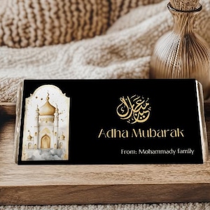Peut inclure: Emballage de barre chocolat&eacute;e noire avec texte dor&eacute; et illustration d'une mosqu&eacute;e. Le texte indique "Adha Mubarak" et "From: Mohammady family". Pr&eacute;sent&eacute; sur un plateau en bois.