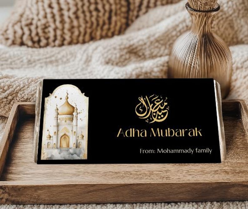 Peut inclure: Emballage de barre chocolat&eacute;e noire avec texte dor&eacute; et illustration d'une mosqu&eacute;e. Le texte indique "Adha Mubarak" et "From: Mohammady family". Pr&eacute;sent&eacute; sur un plateau en bois.