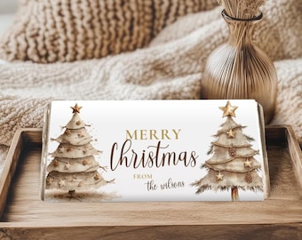 May include: A white rectangular gift box with the words "MERRY Christmas" in gold script, flanked by watercolor Christmas trees. The words "from the wilsons" are below the main text. The box is on a wooden tray.