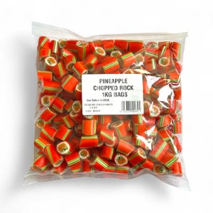 May include: A transparent bag containing pineapple-flavored chopped rock candy. The candy is orange with green stripes and pineapple designs. The label reads "PINEAPPLE CHOPPED ROCK 1KG BAGS".