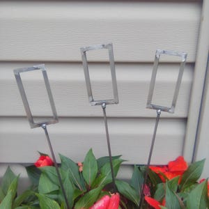 May include: Three silver-colored metal rectangular frames attached to thin metal stakes. Each frame is welded to a vertical rod, designed to be inserted into the ground. The metal has a raw, industrial finish with visible weld points at the joints.