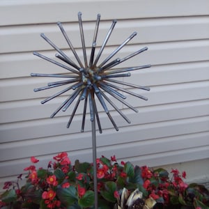 May include: A metal garden sculpture resembling a spiky sphere, with numerous rods radiating outwards. The sculpture is mounted on a slender pole and set amidst vibrant red flowers and green foliage. The sculpture's height is approximately 48 inches.