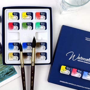 Może przedstawiać: Zestaw farb akwarelowych z różnymi kolorami w białej palecie, w zestawie z dwoma pędzlami i granatowym pudełkiem z napisem "Watercolours".