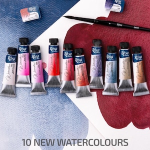 May include: A collection of ten new watercolor paint tubes in various colors, arranged on a white surface with red and blue accents. The tubes are labeled with the brand "Rósa" and color numbers. A paintbrush and two watercolor paint blocks are also present.