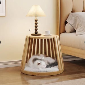 Puede incluir: Una cama para gatos de madera clara con un cojín de piel sintética blanca, una pequeña lámpara de mesa y un gato durmiendo. La lámpara tiene una pantalla plisada y una base de madera. El gato es blanco y gris.