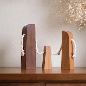 Puede incluir: Tres objetos decorativos de madera de diferentes alturas, conectados por una cuerda blanca. El más alto es marrón oscuro, el del medio es marrón claro y el más corto es marrón más claro. Los objetos están sobre una superficie de madera.