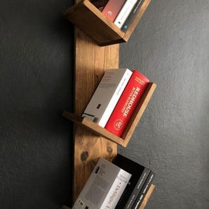 Op de afbeelding: Een rustieke, houten wandplank met drie schuine planken. Elke plank bevat verschillende boeken met zichtbare titels, waaronder "Sapiens" en "Redhouse". De boekenplank is gemaakt van lichtgekleurd hout en is bevestigd aan een donkergrijze muur.
