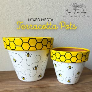 Op de afbeelding: Twee terracotta potten met een bijen thema. De potten zijn wit met gele randen met een zwart honingraatpatroon. Kleine bijen zijn op het witte oppervlak geschilderd, met gestippelde vliegroutes. De tekst "MIXED MEDIA Terracotta Pots" wordt weergegeven.