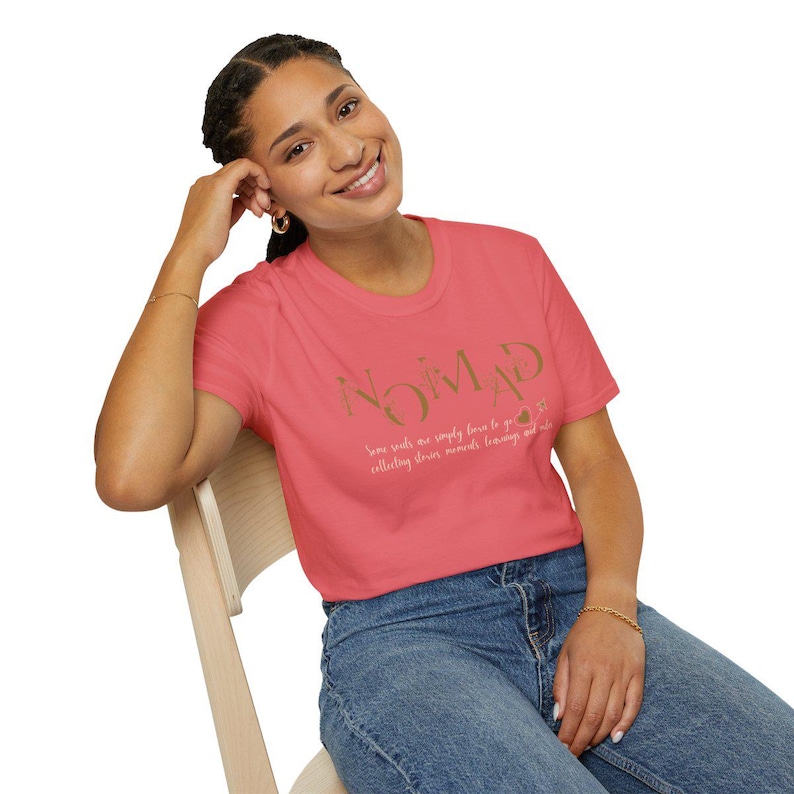 May include: A coral-colored t-shirt with the word "NOMAD" in gold lettering. Below the word is the phrase "Some souls are simply born to go collecting stories, moments, learnings and more." The model is wearing blue jeans.