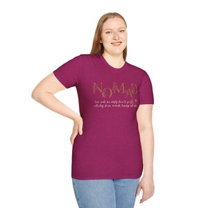 May include: A magenta t-shirt with the word "NOMAD" in gold script. Below the word, the text reads "Some souls are simply born to go collecting stories, moments, learnings and miles."