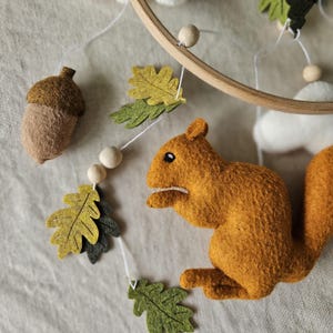 May include: A felt woodland-themed baby mobile featuring a squirrel, acorns, and leaves. The mobile is made from felt and has a wooden ring.