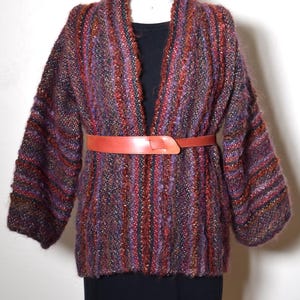 May include: A long-sleeved, open-front cardigan featuring a multi-colored, textured knit pattern in shades of purple, red, and black. The garment is cinched at the waist with a smooth, solid orange belt and is layered over a plain black top.