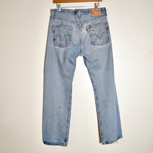 May include: Light blue denim jeans featuring a classic five-pocket design, a tan leather patch on the back waistband, and a small red tab on the right back pocket. The fabric shows signs of wear with light fading and subtle distressing throughout.