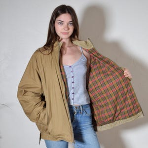 May include: Tan zip-up jacket with a plaid lining. The jacket has a corduroy collar and a relaxed fit. The lining features a red, green, and yellow plaid pattern. The jacket is worn open, revealing a blue and white striped tank top and blue jeans.