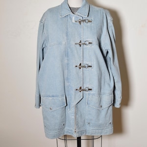 May include: A light blue denim jacket featuring a classic collar, long sleeves, and a front closure with four silver-toned metal toggle clasps. The jacket includes two large flap pockets at the waist and a straight hemline, displayed on a mannequin.