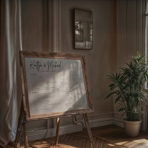 May include: A wooden easel displaying a white crossword puzzle. The puzzle is titled "Kaitlyn & Michael" and features wedding-themed clues. The easel is situated in a room with a mirror and a potted plant.