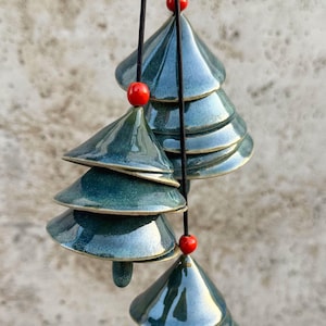 Può includere: Una campanella a vento in ceramica verde acqua con un design a forma di albero di Natale. La campanella a vento presenta più forme di albero impilate, ciascuna con una finitura lucida e un bordo dorato. Perline rosse sono posizionate tra gli alberi. Appesa a un filo nero.
