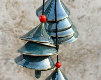 Op de afbeelding: Een hangende teal keramische windgong met een kerstboomontwerp. De windgong heeft meerdere gestapelde boomvormen, elk met een glanzende afwerking en een gouden rand. Rode kralen zijn tussen de bomen geplaatst. De windgong hangt aan een zwarte draad.